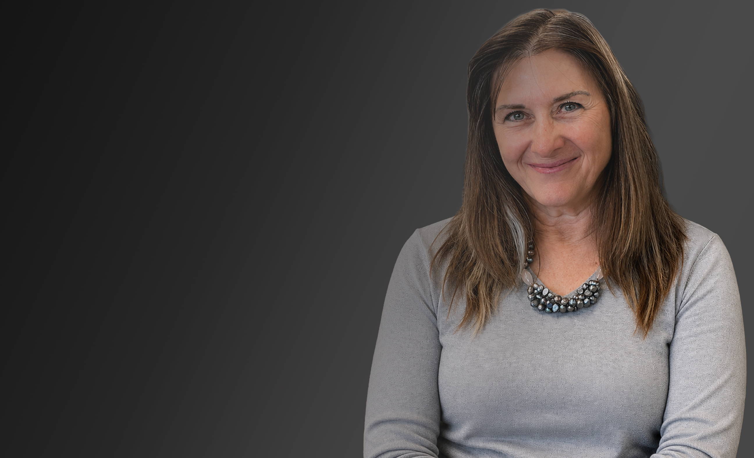 A woman with shoulder-length brown hair smiling, wearing a light gray long-sleeve top and a dark beaded necklace, set against a dark gray gradient background.